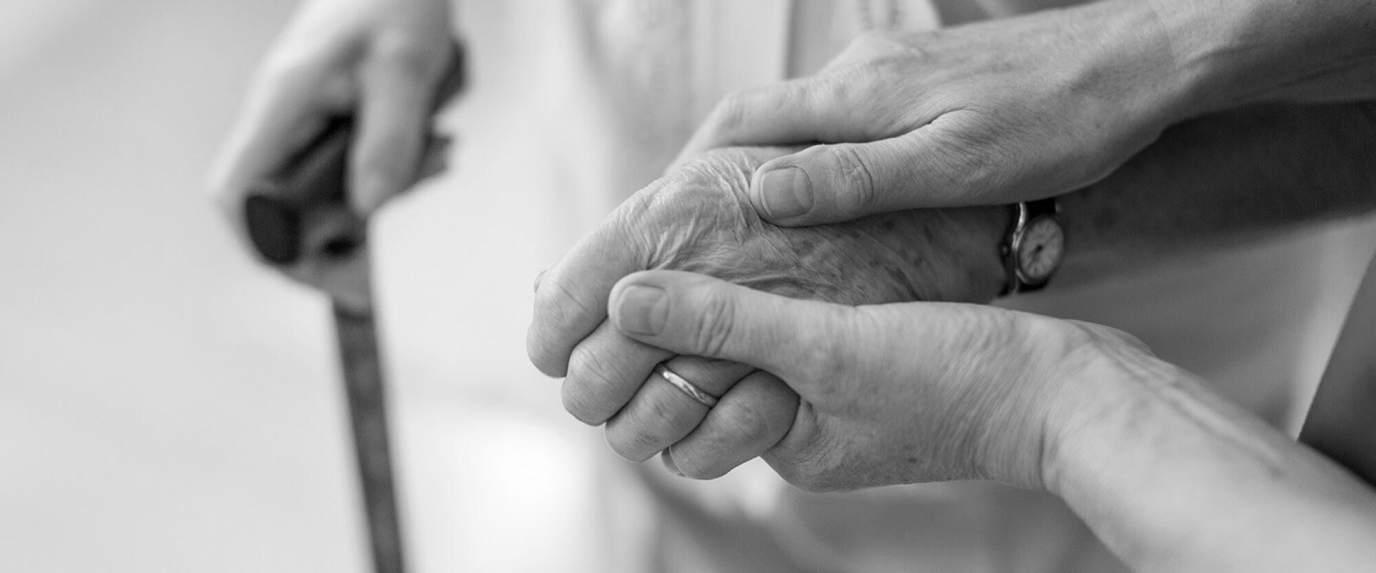 An older person holding a walking stick with a supporting hand resting over theirs