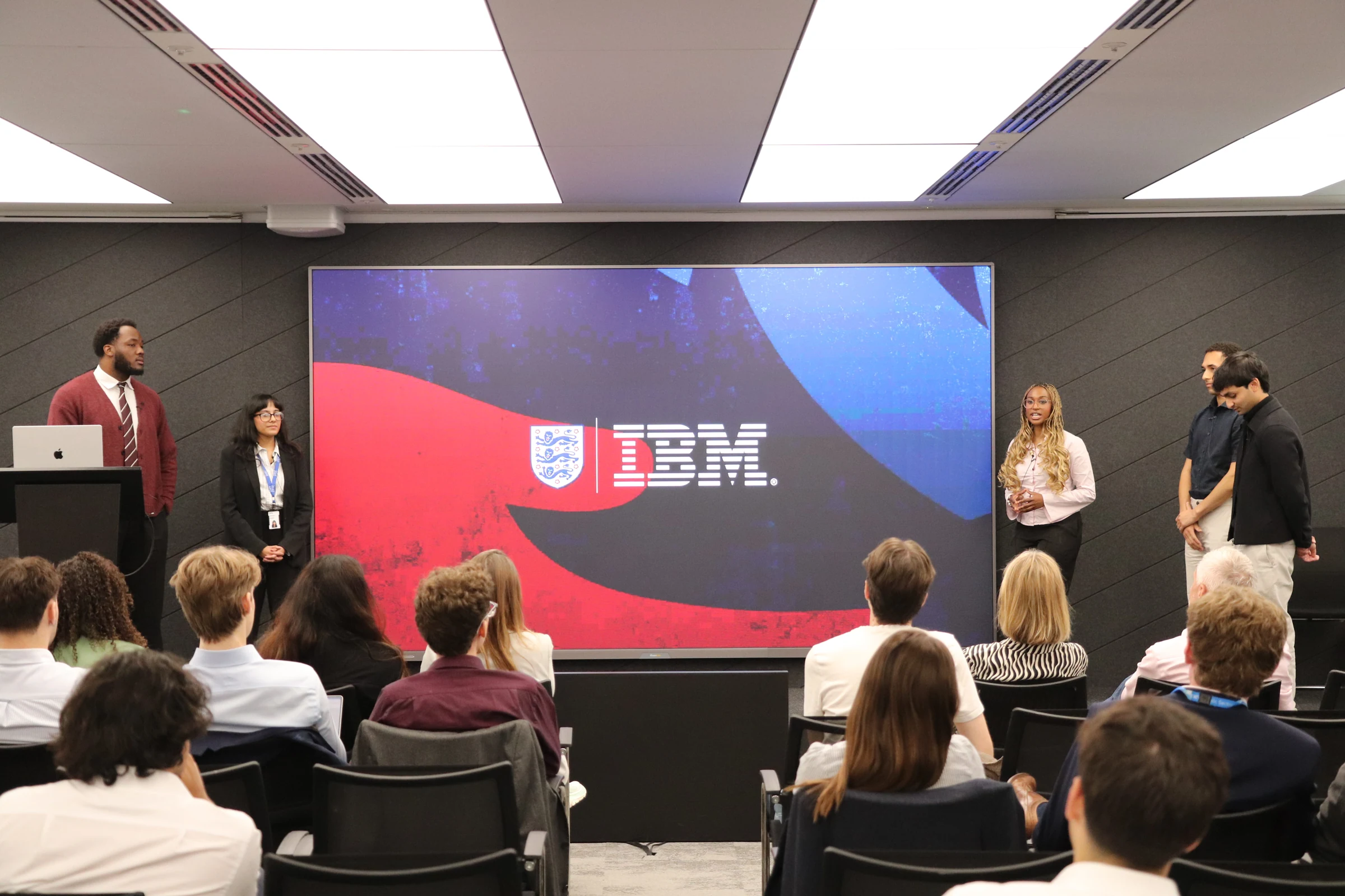 Yusuf and the internship team presenting beside a large FA and IBM display at the showcase