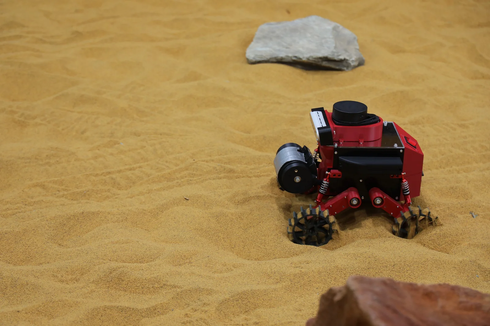 Olympus Advanced rover in sand beside rocks.