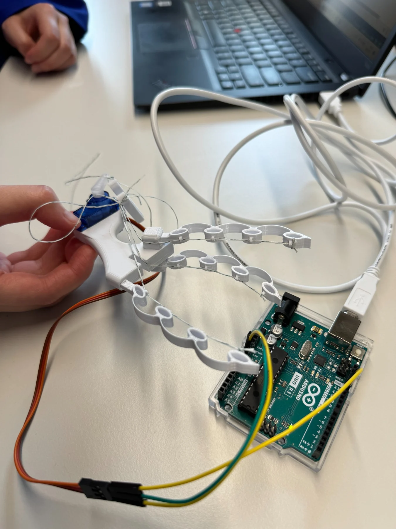 White compliant gripper with strings, servo and Arduino on a workbench.